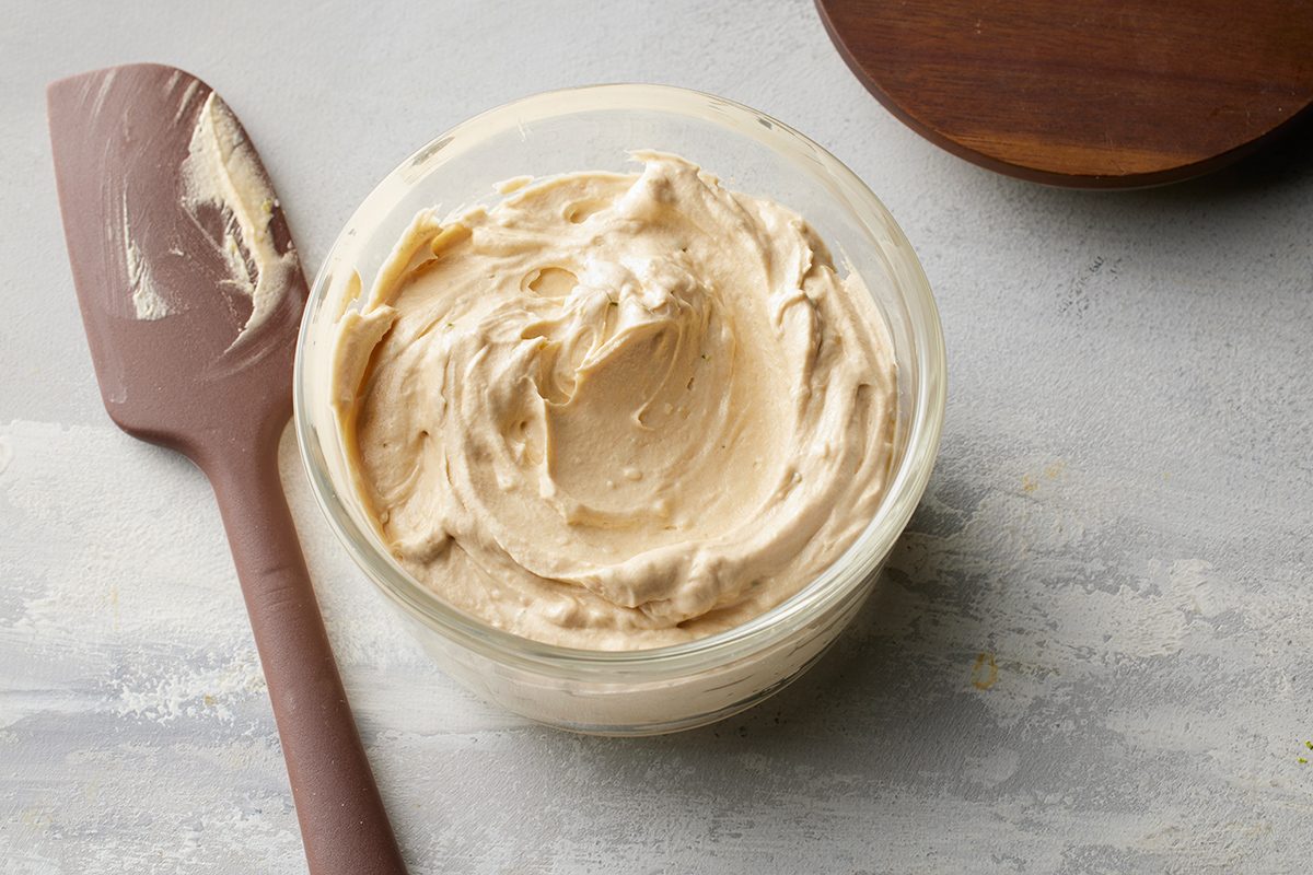 A glass bowl filled with creamy, light beige peanut butter sits next to a brown spatula with some peanut butter on it, on a light gray surface.