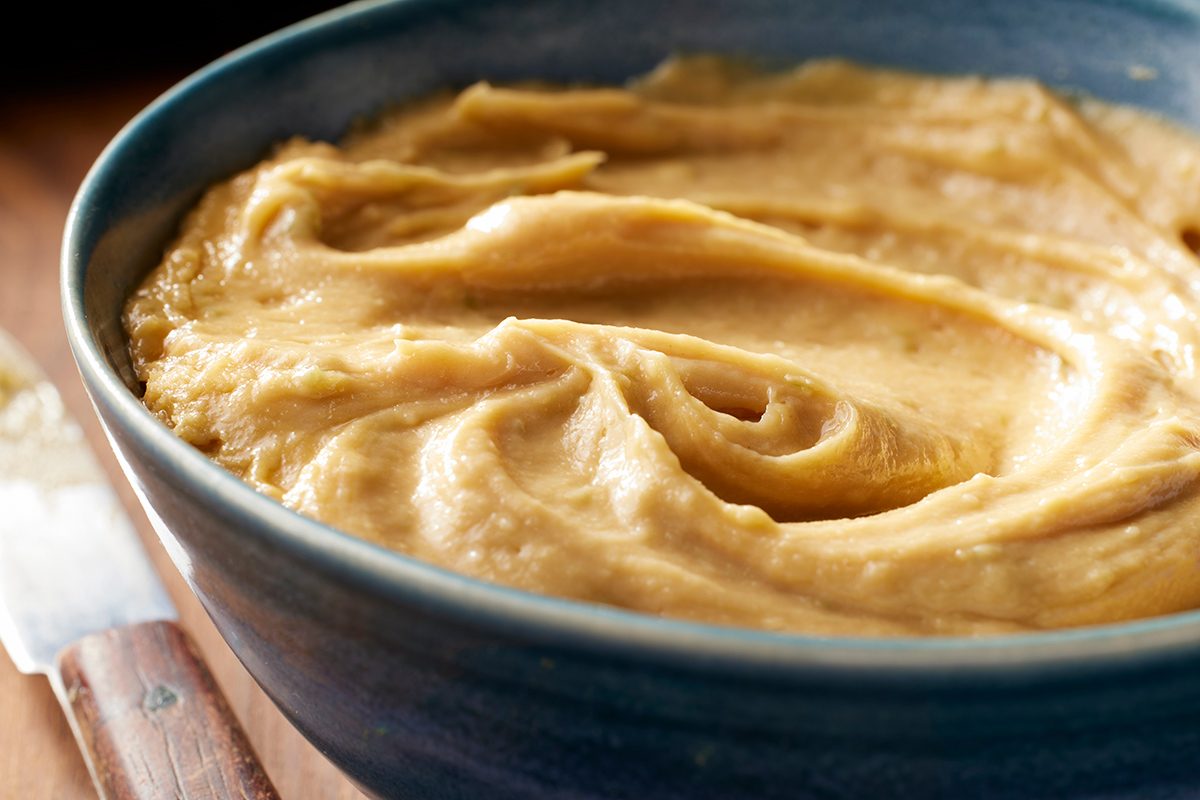A close-up of a blue bowl filled with smooth, creamy peanut butter, with swirls on the surface. A butter knife with a wooden handle is partially visible beside the bowl.