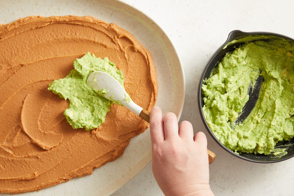 avocado mixture being spread over bean dip base in a serving plate