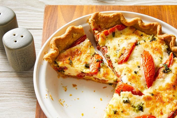 Closeup shot of a basil tomato tart with a slice removed, showing its golden crust, roasted tomatoes, and herb-speckled cheesy filling; Salt and pepper shakers sit nearby on the wooden board