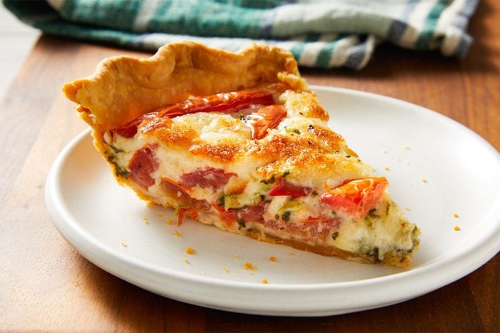 Closeup shot of a single slice of basil tomato tart on a white plate, showing its flaky crust, roasted tomatoes, and creamy, herb-filled interior; A striped kitchen towel sits in the background;