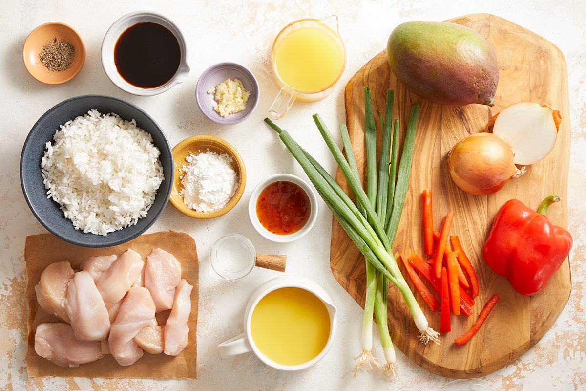 Overhead shot of tropical chicken ingredients laid out on a cutting board and countertop, including diced chicken, bell peppers, onion, green onion, mango, rice, and assorted sauces in small bowls.