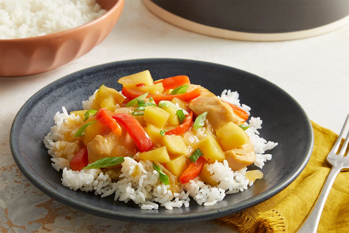 3/4 shot of plated Tangy Tropical Chicken served over a bed of white rice on a black dish, garnished with green onion slices.