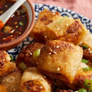 Close-up of crispy golden spring rolls garnished with sesame seeds and chopped green onions, served on a patterned plate with a small bowl of dipping sauce on the side.