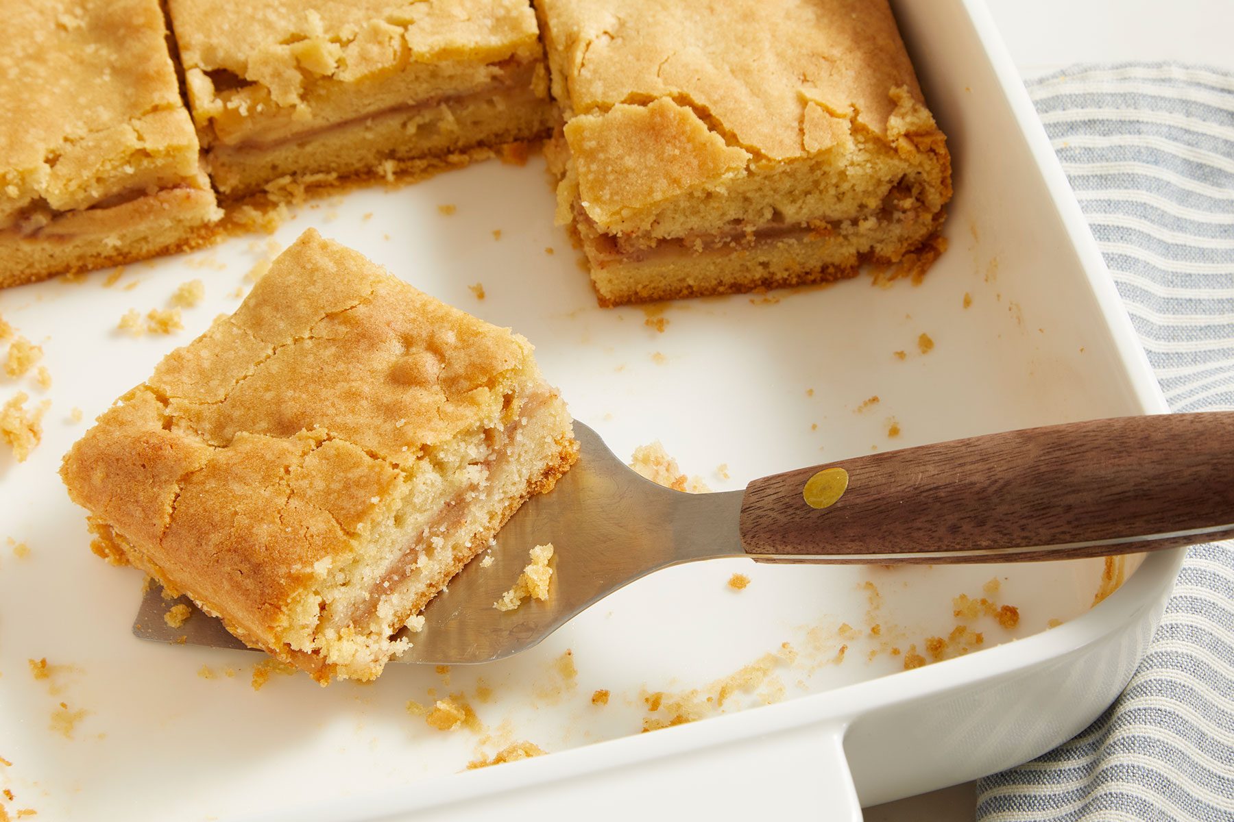 Small pieces of Apple Cake in a white colored dish with one piece being lifted to serve by a serving knife.
