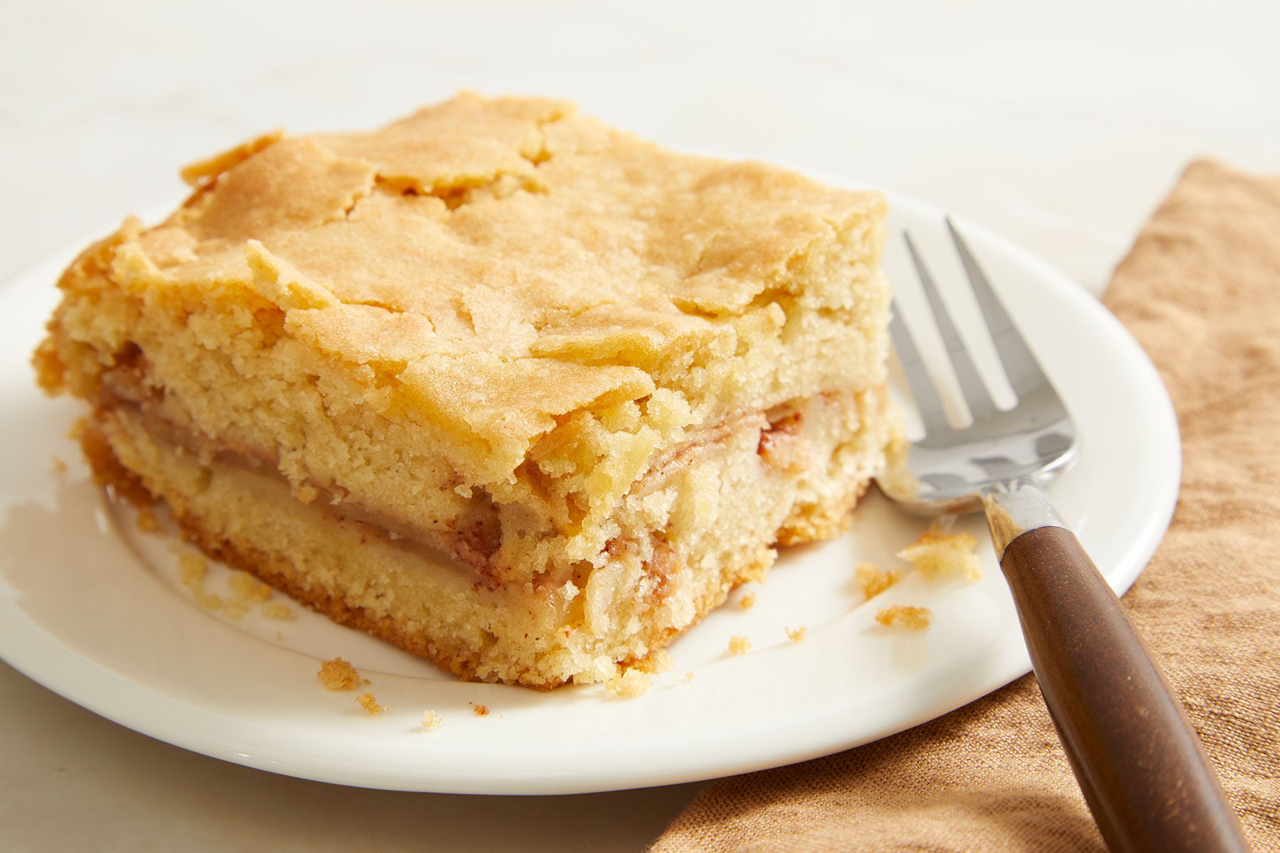 A small piece of Apple Cake placed on a white colored plate with a fork placed on side.