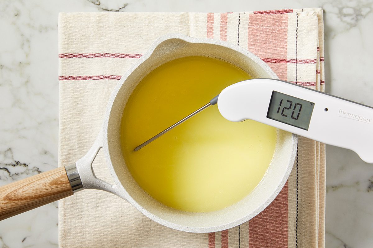 A saucepan filled with melted yellow liquid is placed on a striped kitchen towel. A digital thermometer inserted into the liquid displays a temperature of 120 degrees.