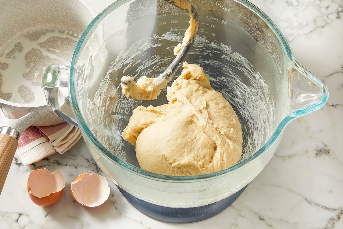 A glass mixing bowl with cookie dough and a paddle attachment sits on a marble countertop. Next to the bowl are two cracked eggshells and a saucepan with a cloth underneath.