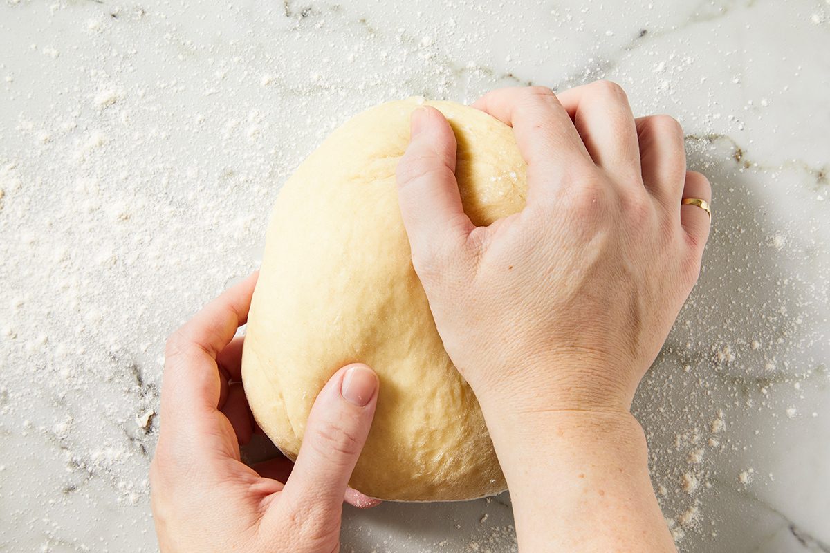 Two hands kneading a ball of dough on a floured, light-colored marble surface.