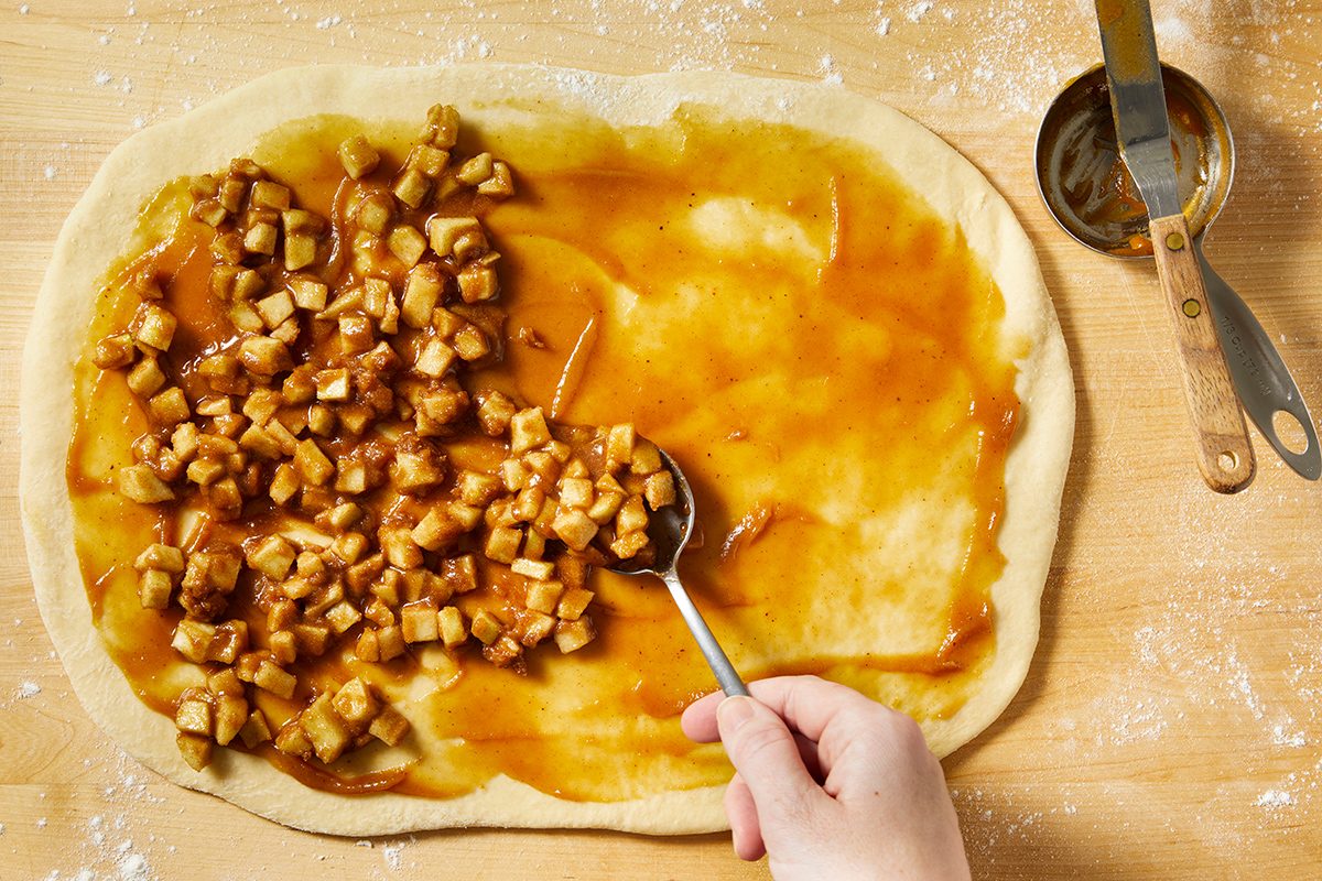 A hand spreads cinnamon-coated diced apples and syrup over rolled-out dough on a floured wooden surface, with a measuring cup and spoon nearby.