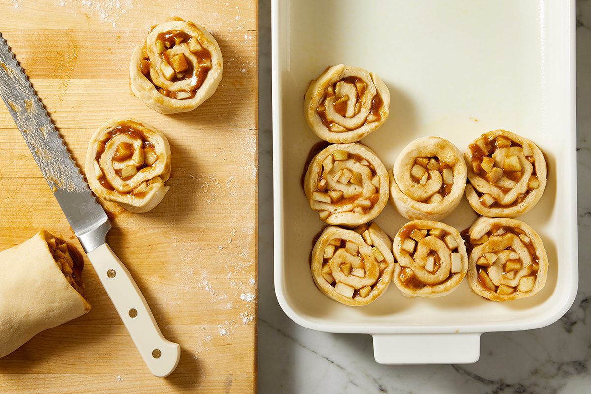 Unbaked cinnamon rolls with visible apple pieces are arranged in a white baking dish; others are on a wooden board beside a serrated knife. The scene is set on a marble countertop.