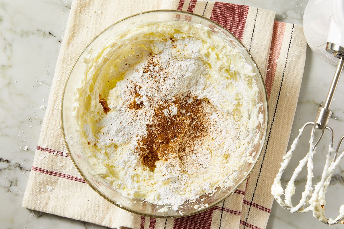 A glass mixing bowl with creamy batter, powdered sugar, and cinnamon on top, sits on a striped cloth. An electric mixer with beaters covered in batter rests nearby on a marble countertop.