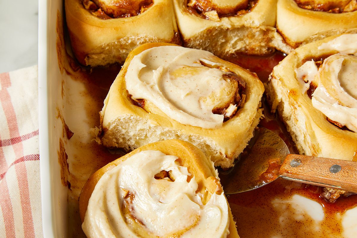 A baking dish filled with freshly baked cinnamon rolls topped with icing; one roll is being lifted out with a spatula.