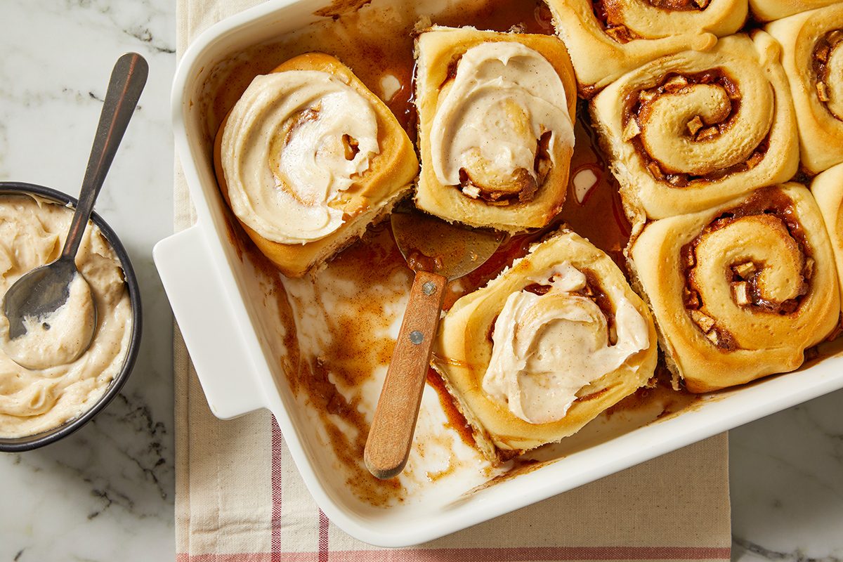 A baking dish filled with iced cinnamon rolls, some with frosting on top, sits on a table next to a small bowl of frosting and a spoon. A spatula rests in the dish where one roll has been removed.