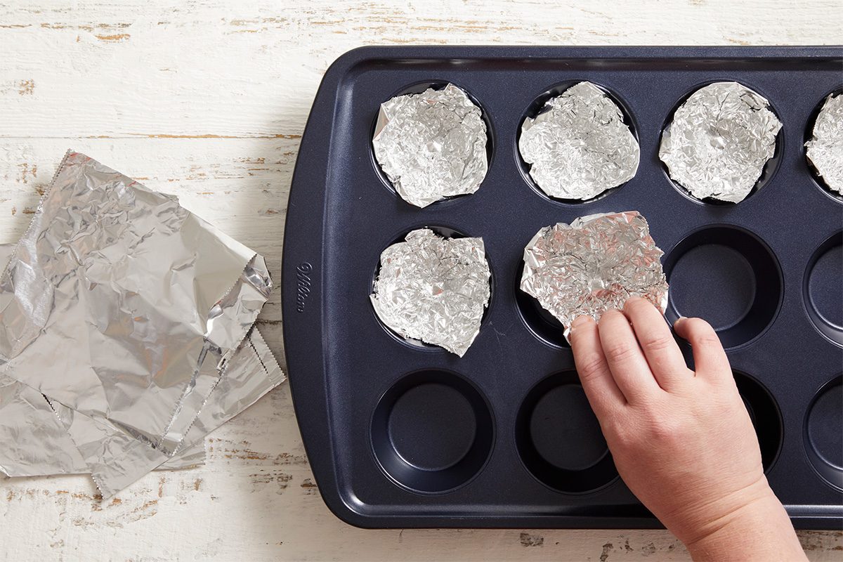 Overhead shot of a muffin pan lined with circular molds made of aluminum foil, a hand pressing foil into shape.