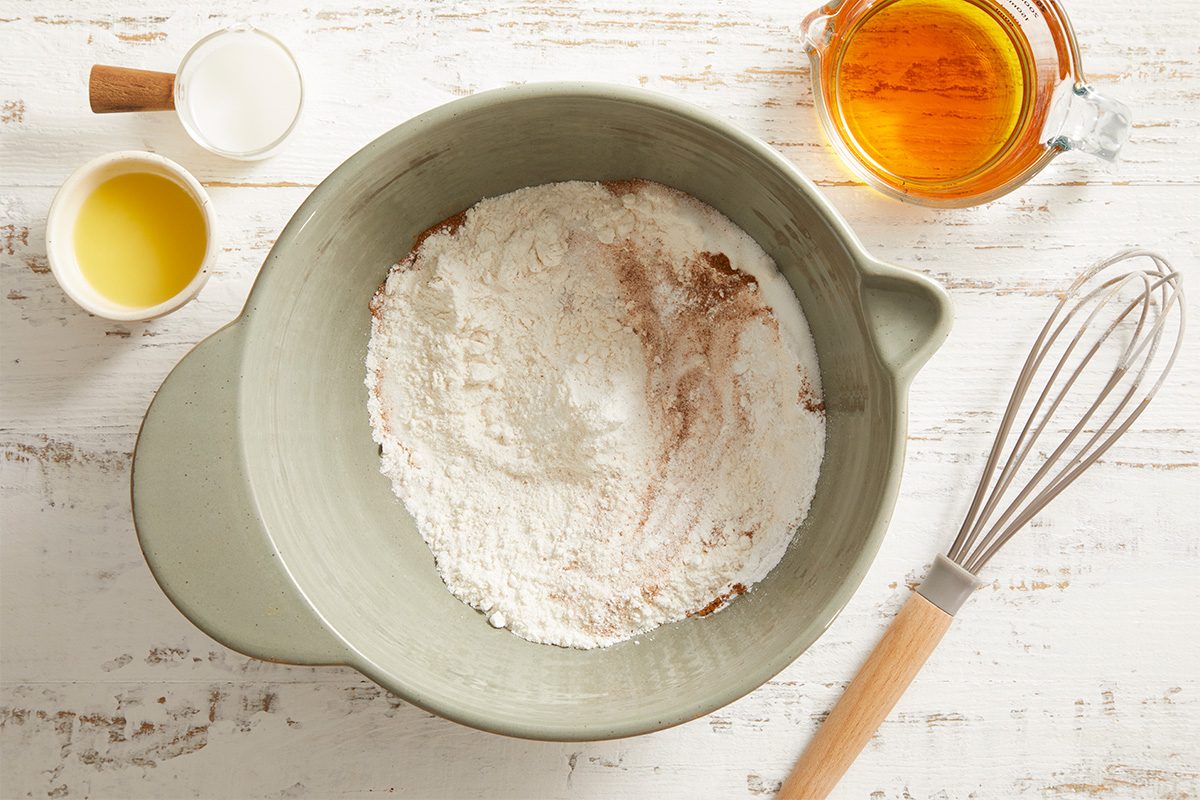 Overhead shot of a mixing bowl containing flour, sugar, and a whisk, with honey and an egg placed nearby.
