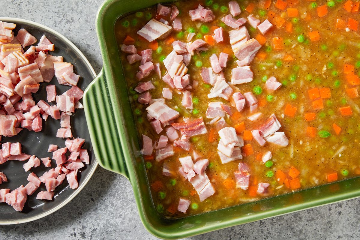 Overhead step-by-step preparation image showing diced ham being added to a green ceramic baking dish filled with uncooked long-grain rice, chicken broth, soy sauce, peas, carrots, edamame, and seasonings. The ingredients are combined to prepare baked fried rice before cooking, with chopped bacon pieces visible nearby on a gray countertop.
