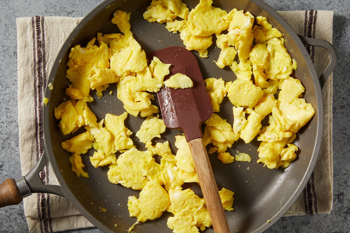 Overhead step-by-step cooking image showing scrambled eggs being cooked in a nonstick skillet using a silicone spatula. The eggs are lightly scrambled as a component that will be folded into the baked fried rice dish.