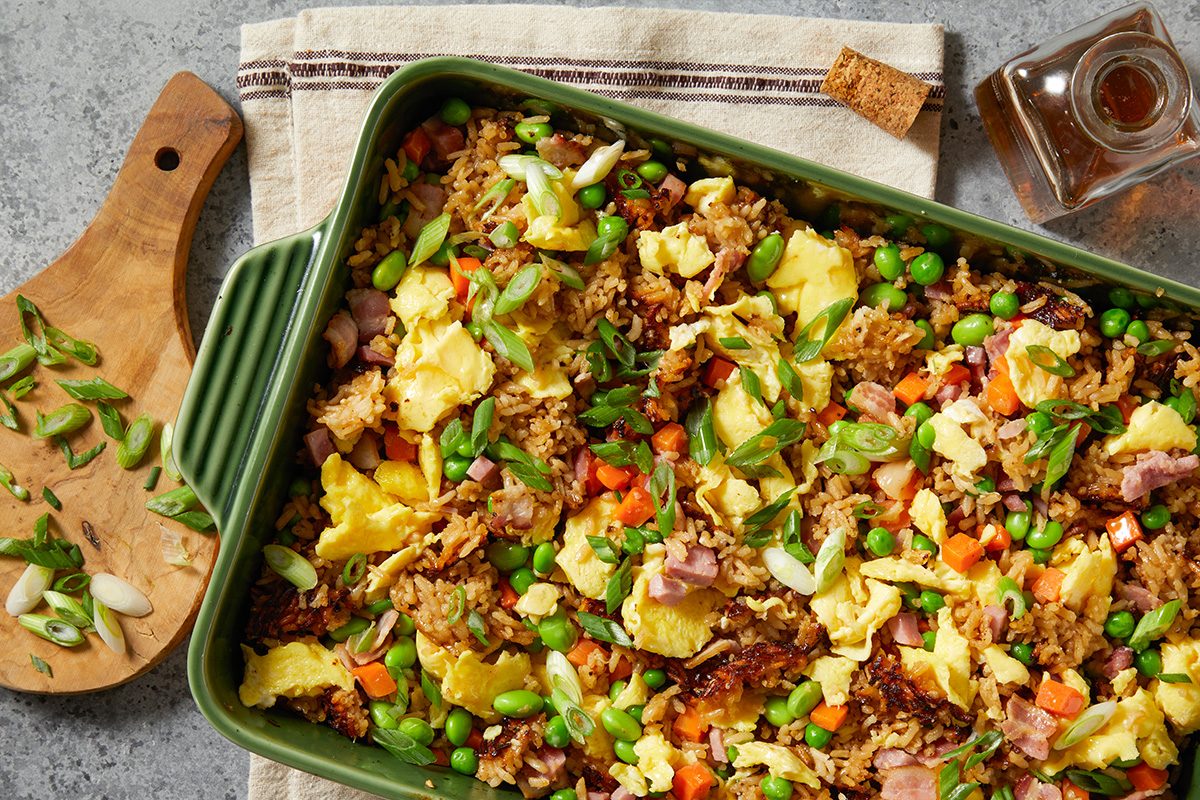 Overhead step-by-step image showing baked fried rice after cooking in a green ceramic baking dish. The dish is topped with scrambled eggs, diced ham, bacon pieces, peas, carrots, edamame, and sliced green onions, creating a colorful baked version of classic fried rice.