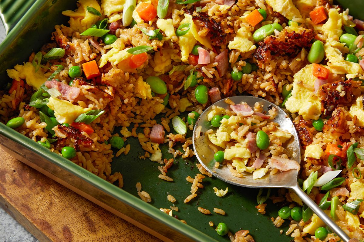 Close-up overhead push-and-pull image showing a serving spoon scooping baked fried rice from a green ceramic casserole dish. The spoon reveals a mixture of fluffy rice, scrambled eggs, diced ham, crispy bacon pieces, peas, carrots, edamame, and green onions.