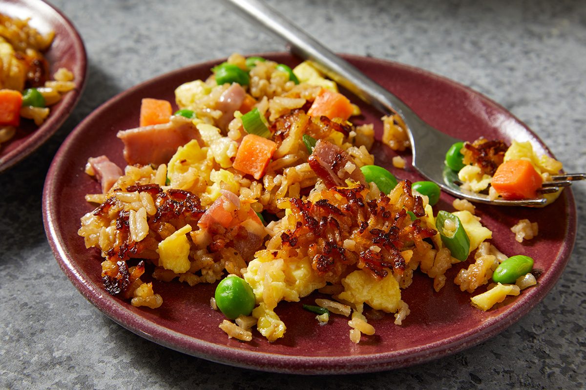 Push-and-pull plated serving image showing a portion of baked fried rice served on a red plate. The rice is topped with scrambled eggs, diced ham, bacon, peas, carrots, and edamame, with a fork lifting a bite from the dish.