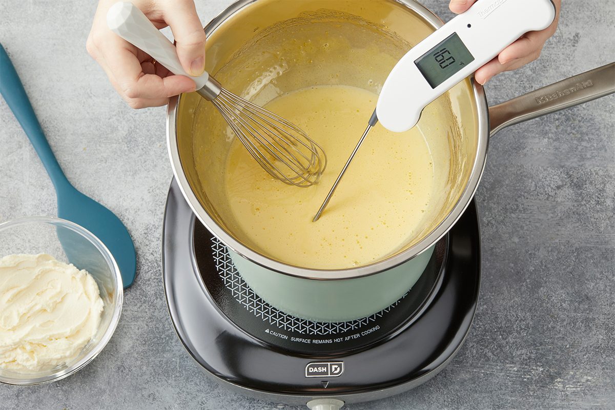 Whisk egg yolks, 1/2 cup sugar, 2 tablespoons banana liqueur and salt on top of a double boiler until mixture is thickened (ribbon stage) and a thermometer reads 160°. Overhead premium food photograph of egg yolks, sugar, and salt being whisked over gentle heat to create the custard base for Banana Pudding Tiramisu. A thermometer is visible for precision temperature control.