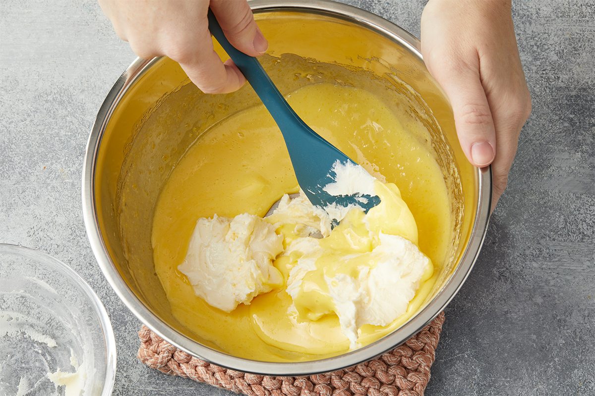 Remove from heat; stir in one tub of mascarpone cheese and banana extract until smooth. Overhead view of mascarpone cheese being folded into the warm custard mixture to create a smooth and creamy filling for Banana Pudding Tiramisu.