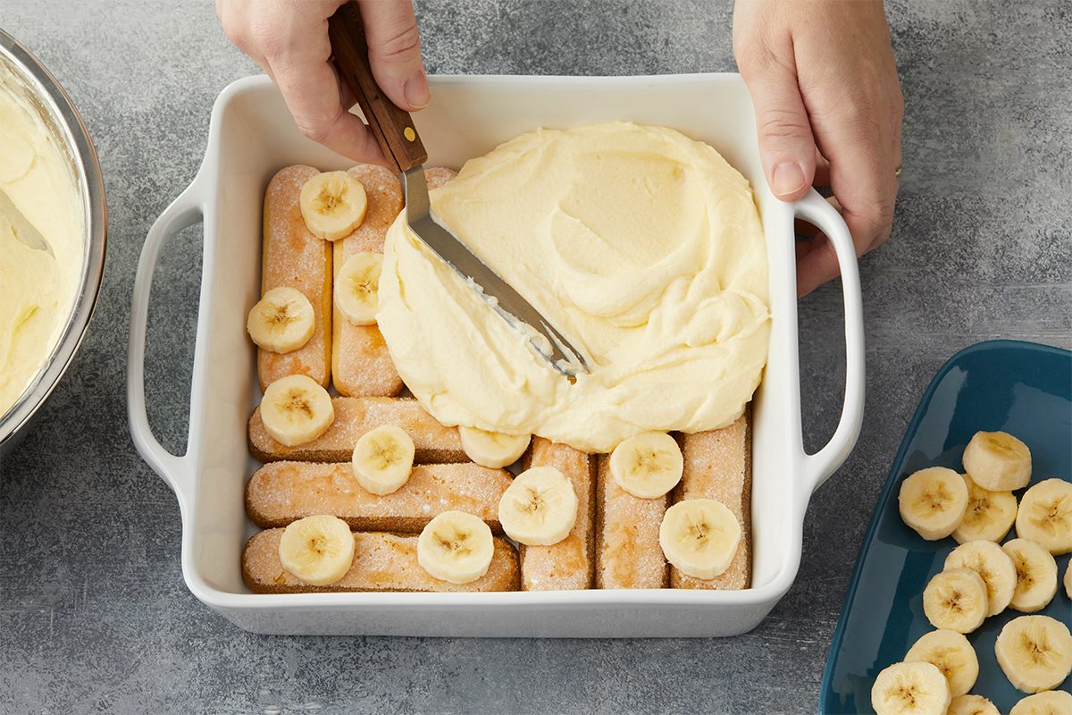 Top with half the banana slices. Spread half the mascarpone mixture over top. Overhead image of soaked ladyfingers layered with sliced bananas and creamy mascarpone filling in a baking dish during assembly of Banana Pudding Tiramisu.