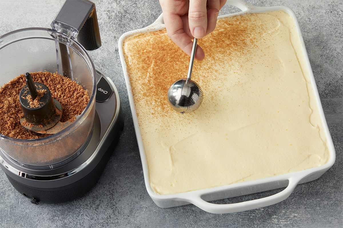 In a food processor; blitz banana chips into a fine powder. Add cocoa until combines. Dust banana cocoa over tiramisu before serving. Overhead premium photograph of baking cocoa being sifted over the top layer of mascarpone filling to finish the Banana Pudding Tiramisu before chilling.