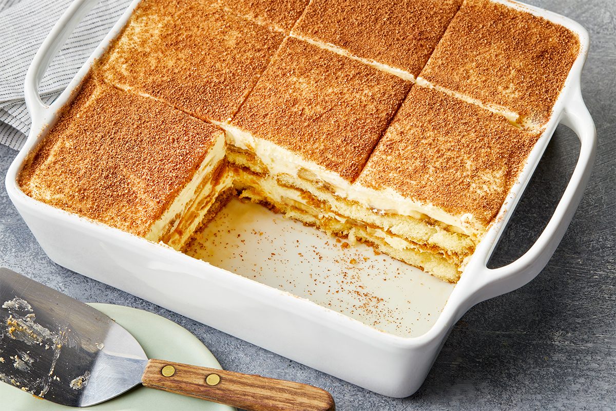 Overhead premium photograph of the full Banana Pudding Tiramisu in a white baking dish with one square removed to reveal the layered interior.