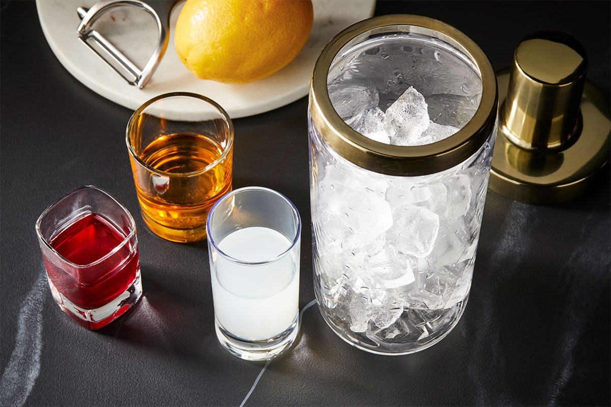 Overhead horizontal preparation image for Belmont Jewel (ID: 282195) featuring ice cubes in a glass ice bucket alongside measured bourbon, lemonade and pomegranate juice with fresh lemon. Premium set photography styled for Taste of Home, capturing the step-by-step cocktail setup before mixing.