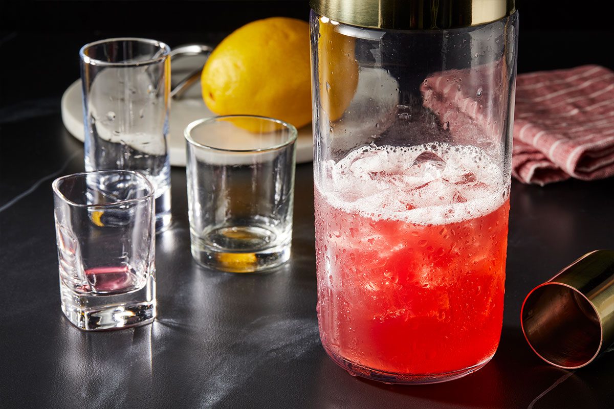 Horizontal overhead image of a cocktail shaker filled with ice, bourbon, lemonade and pomegranate juice being mixed for the Belmont Jewel (ID: 282195). Premium set step-by-step photography styled for Taste of Home highlighting vibrant red color and cocktail preparation process.