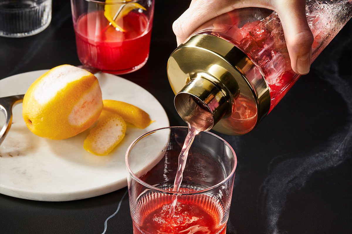 Horizontal action shot of Belmont Jewel (ID: 282195) being strained from a cocktail shaker into a rocks glass filled with ice. Premium set push-and-pulls image styled for Taste of Home capturing the rich ruby color and smooth pour.