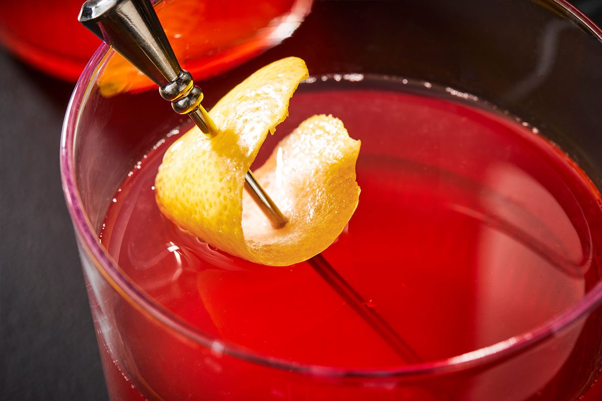 Horizontal close up image of Belmont Jewel (ID: 282195) highlighting lemon peel garnish suspended over the deep red cocktail surface. Premium set push-and-pulls photography styled for Taste of Home capturing texture, color and refined cocktail detail.