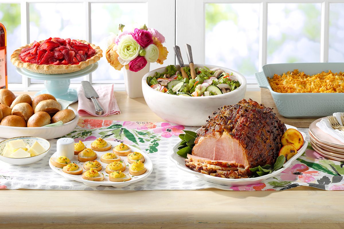 A table set with a glazed ham, deviled eggs, dinner rolls, butter, salad, cheesy casserole, and a strawberry pie, arranged on a floral table runner in front of a bright window.