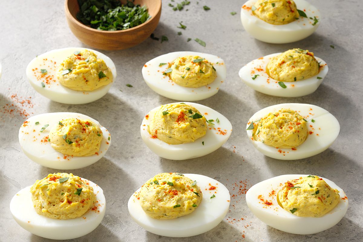 A tray of deviled eggs garnished with herbs and paprika is arranged on a light surface. A small wooden bowl of chopped herbs sits nearby.