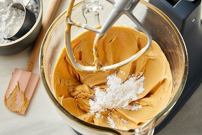 A mixing bowl with creamy peanut butter and powdered sugar being blended with a stand mixer. A spatula with peanut butter and a bowl of powdered sugar are nearby on the counter.