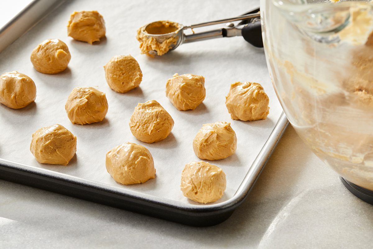A baking tray lined with parchment paper holds several evenly spaced scoops of raw cookie dough, with a cookie scoop and a mixing bowl nearby.