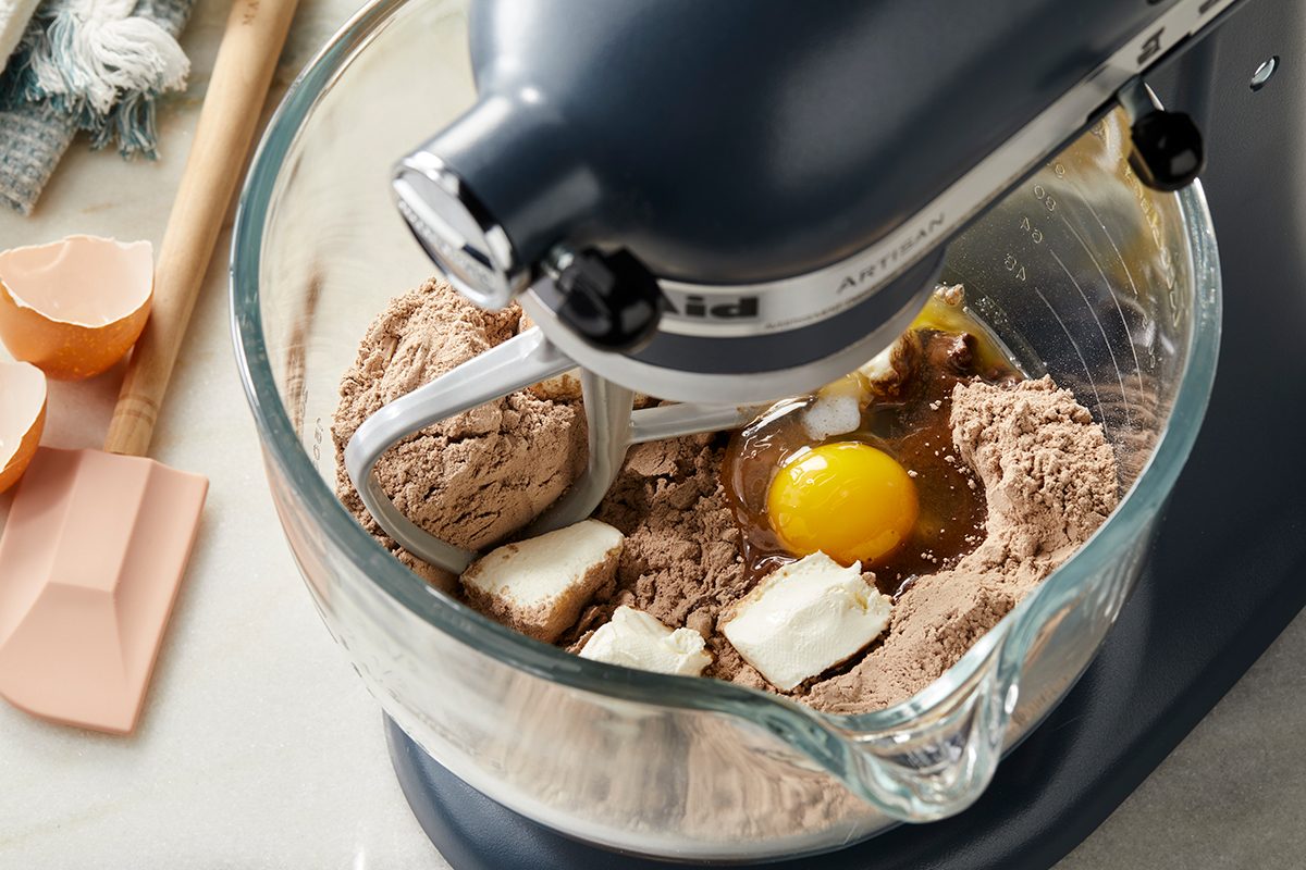 A stand mixer with a glass bowl contains cocoa powder, an egg, and chunks of butter, ready to be mixed. Broken eggshells and a spatula are on the counter nearby.
