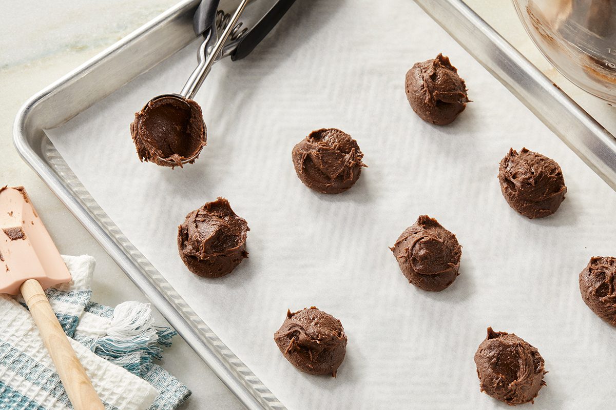 Scoop of chocolate cookie dough on a baking sheet lined with parchment paper, with several dough balls spaced apart, a cookie scoop, spatula, and kitchen towels nearby.