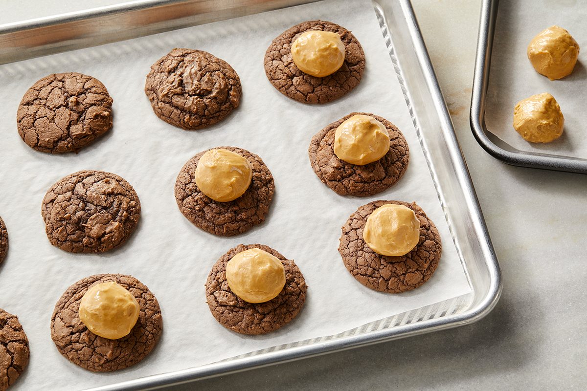 A baking sheet with chocolate cookies, some topped with a round scoop of peanut butter filling. Another tray in the background holds more peanut butter scoops. Both trays are lined with parchment paper.