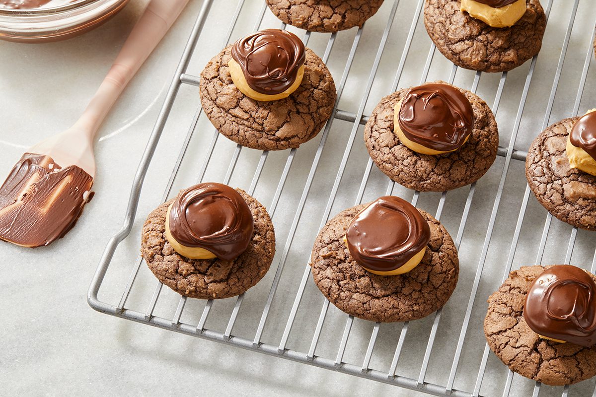 Chocolate cookies with peanut butter and swirled chocolate topping are cooling on a wire rack; a spatula with chocolate is nearby on a light countertop.