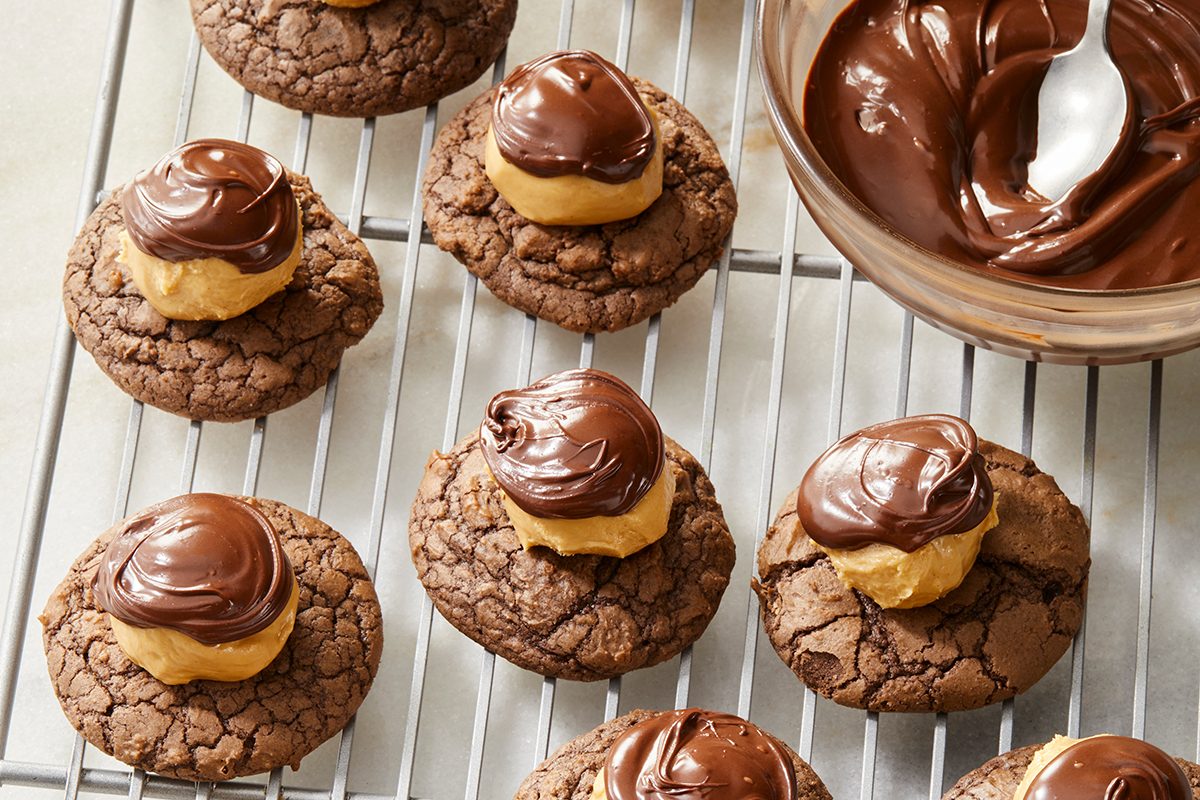 Chocolate cookies topped with a dollop of peanut butter and a swirl of melted chocolate are arranged on a cooling rack. A bowl of melted chocolate with a spoon is beside the cookies.