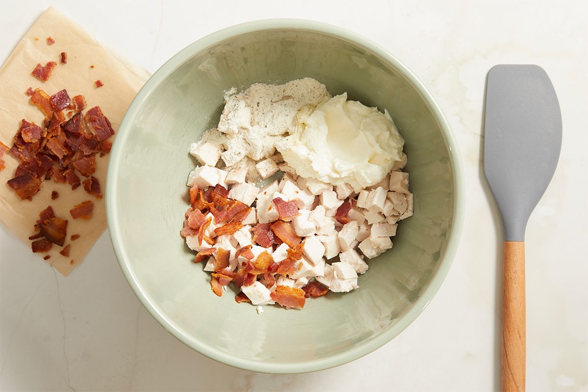 Overhead horizontal step-by-step preparation image showing a bowl of cubed cooked chicken, ricotta cheese, crumbled bacon, and ranch seasoning beside a spatula on a light countertop. Ingredients are ready to be mixed for chicken bacon ranch rice paper spirals.