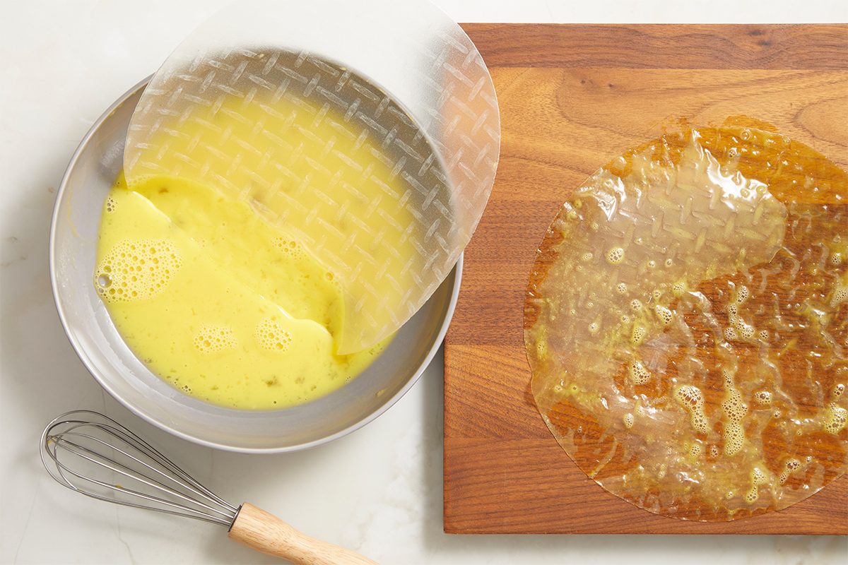 Horizontal overhead step-by-step image showing softened rice paper sheets on a wooden cutting board beside a bowl of beaten eggs and a whisk, preparing wrappers for chicken bacon ranch rice paper spirals.