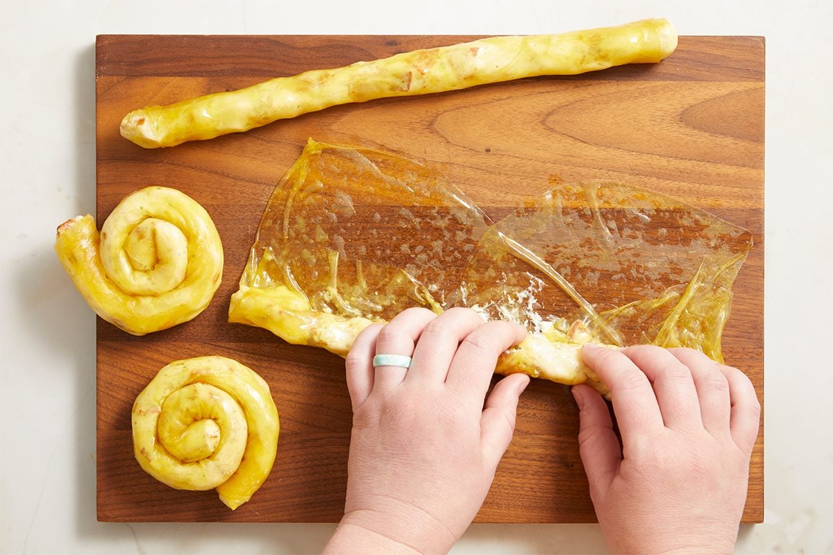 Horizontal overhead step-by-step image showing hands rolling a filled rice paper sheet into spiral shapes on a wooden cutting board to form chicken bacon ranch rice paper spirals.