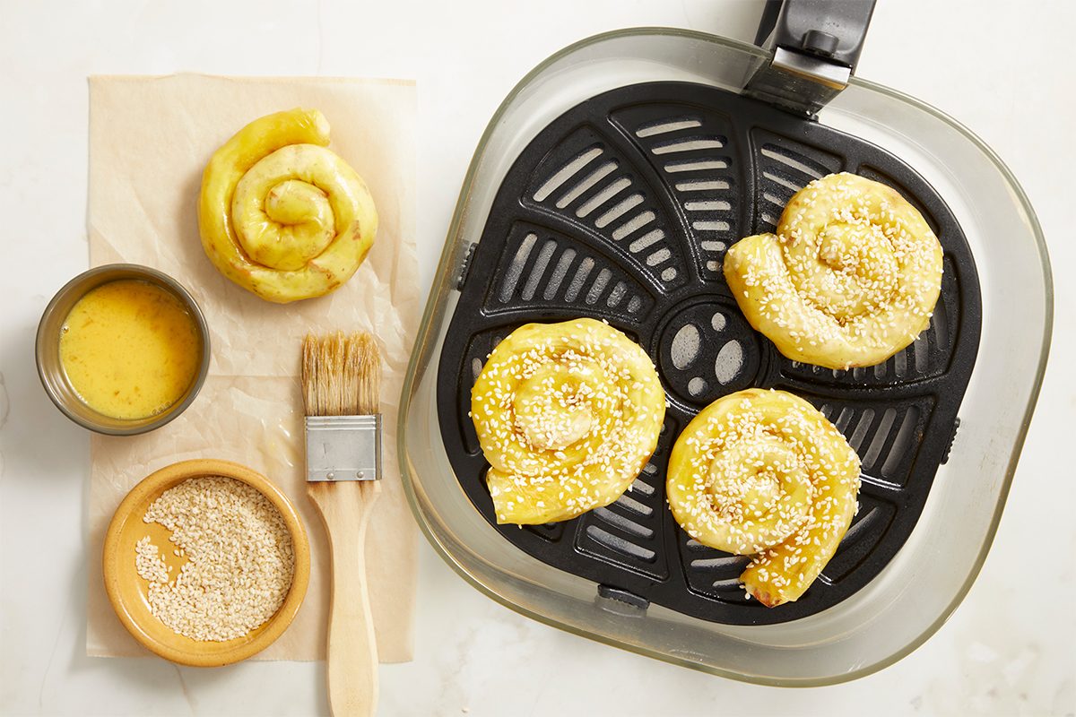 Horizontal step-by-step image showing uncooked sesame-topped chicken bacon ranch rice paper spirals arranged inside an air fryer basket with ranch dressing and sesame seeds nearby.