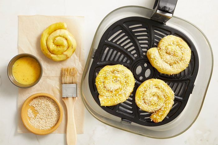 Horizontal step-by-step image showing uncooked sesame-topped chicken bacon ranch rice paper spirals arranged inside an air fryer basket with ranch dressing and sesame seeds nearby.