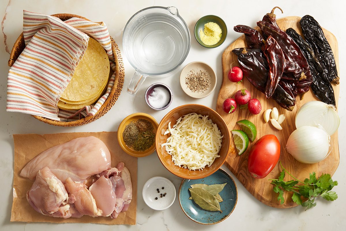 Ingredients for a Mexican dish arranged on a counter, including chicken breasts, tortillas, cheese, dried chiles, tomato, onion, garlic, radishes, herbs, butter, spices, water, and a lime wedge.