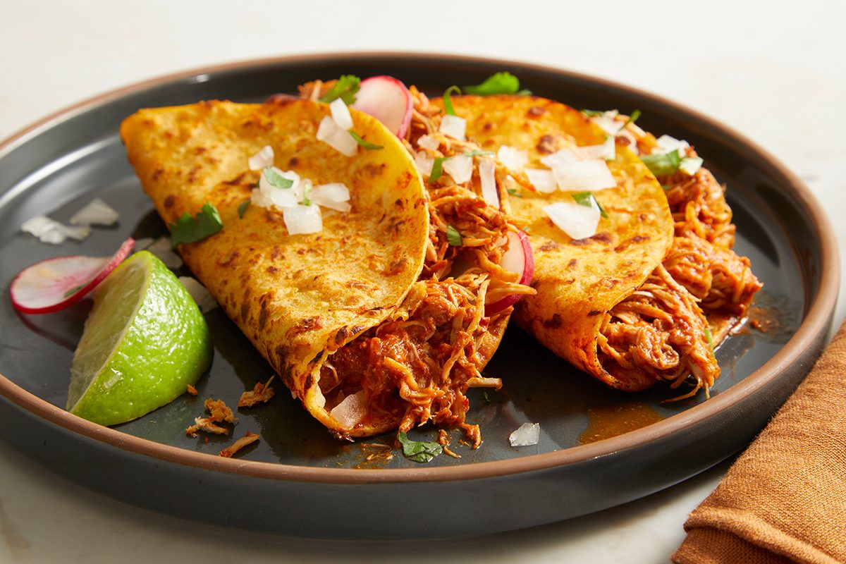 Two crispy tacos filled with shredded meat, topped with chopped onions and cilantro, garnished with sliced radishes and a lime wedge, served on a dark plate.